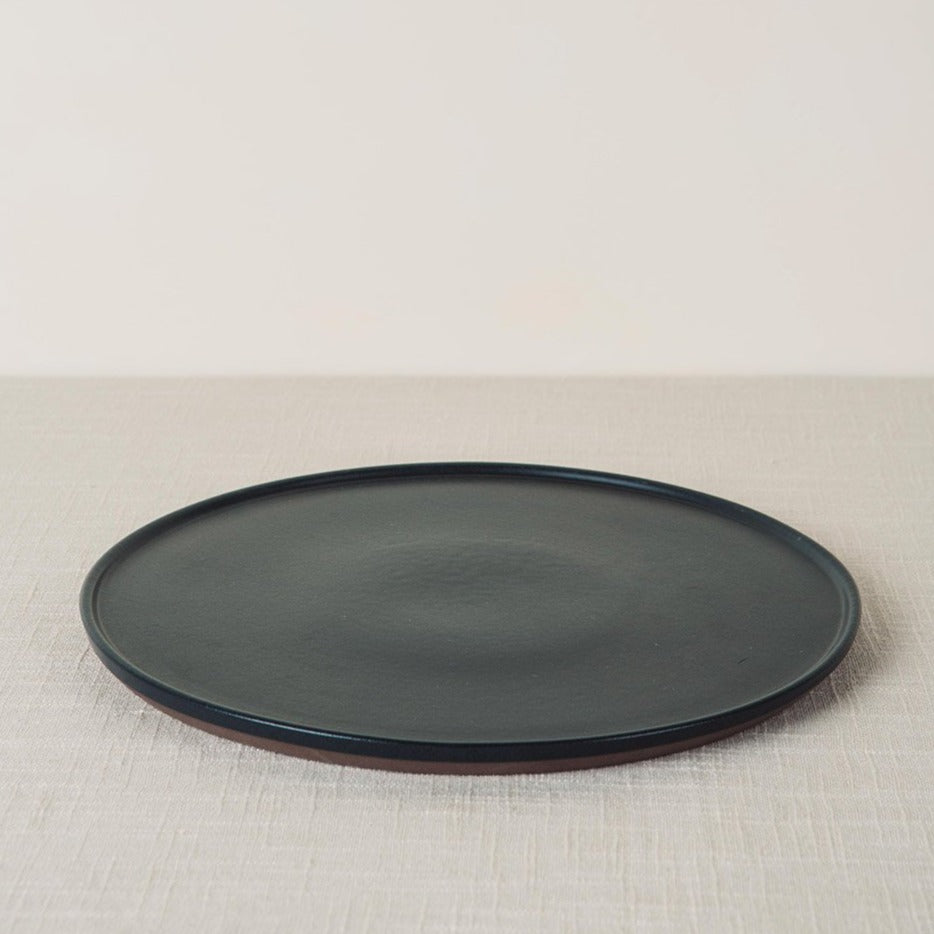 Onyx dinner plate with a dark matte surface and reddish-brown rim, angled on a light textured surface.