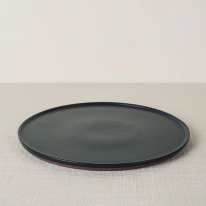 Onyx dinner plate with a dark matte surface and reddish-brown rim, angled on a light textured surface.