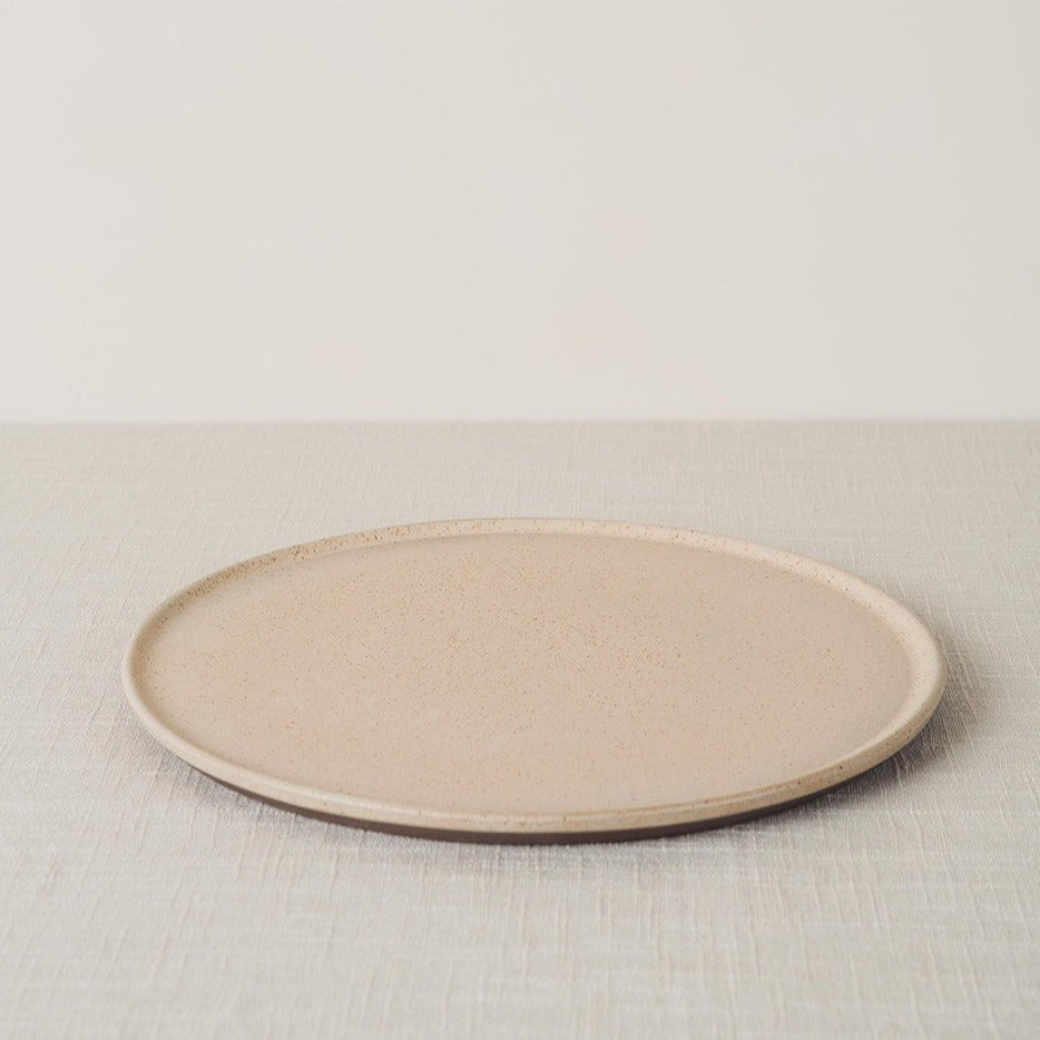 Light beige sandstone dinner plate with a speckled finish and dark exposed stoneware rim, angled on a linen surface.