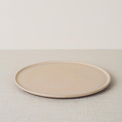 Light beige sandstone dinner plate with a speckled finish and dark exposed stoneware rim, angled on a linen surface.