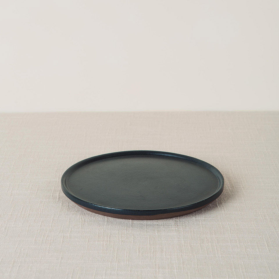 Dark onyx side plate with a matte finish and unglazed brown edge, placed on a beige linen surface.
