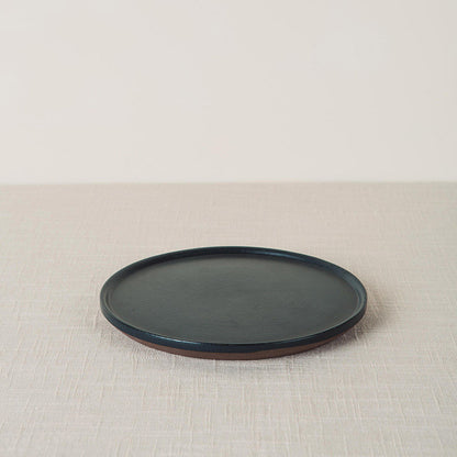 Dark onyx side plate with a matte finish and unglazed brown edge, placed on a beige linen surface.