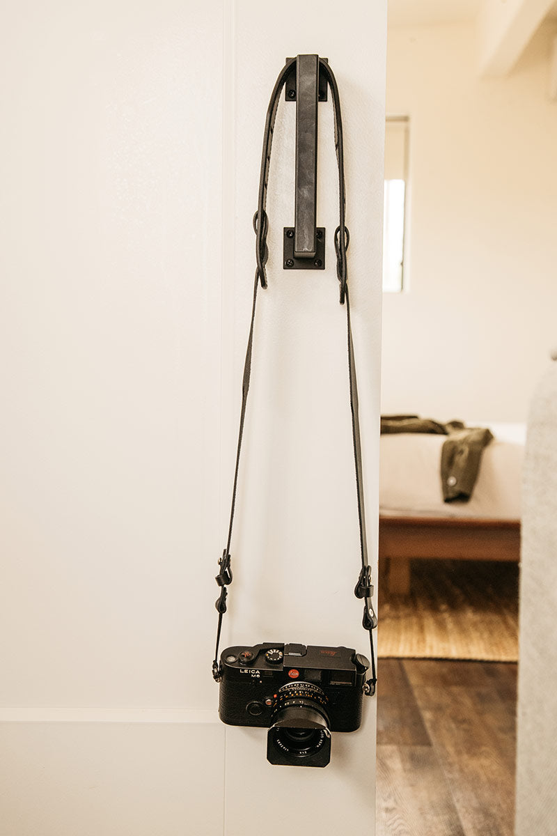 Black SLR camera strap hanging from a dark metal hook, with a black vintage-style camera attached.