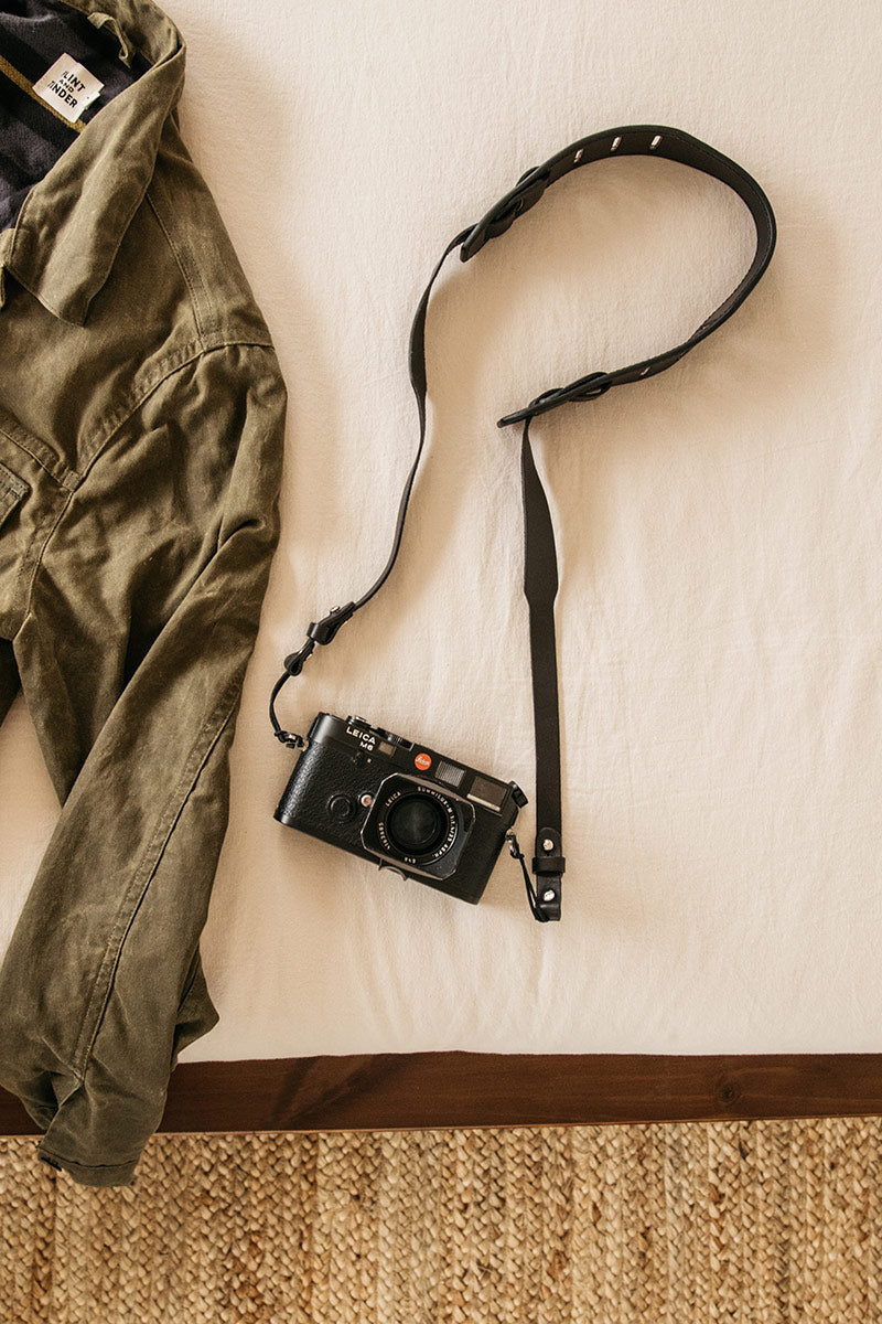 Black leather SLR camera strap with adjustable stud details, attached to a camera, next to a green jacket on a white sheet.