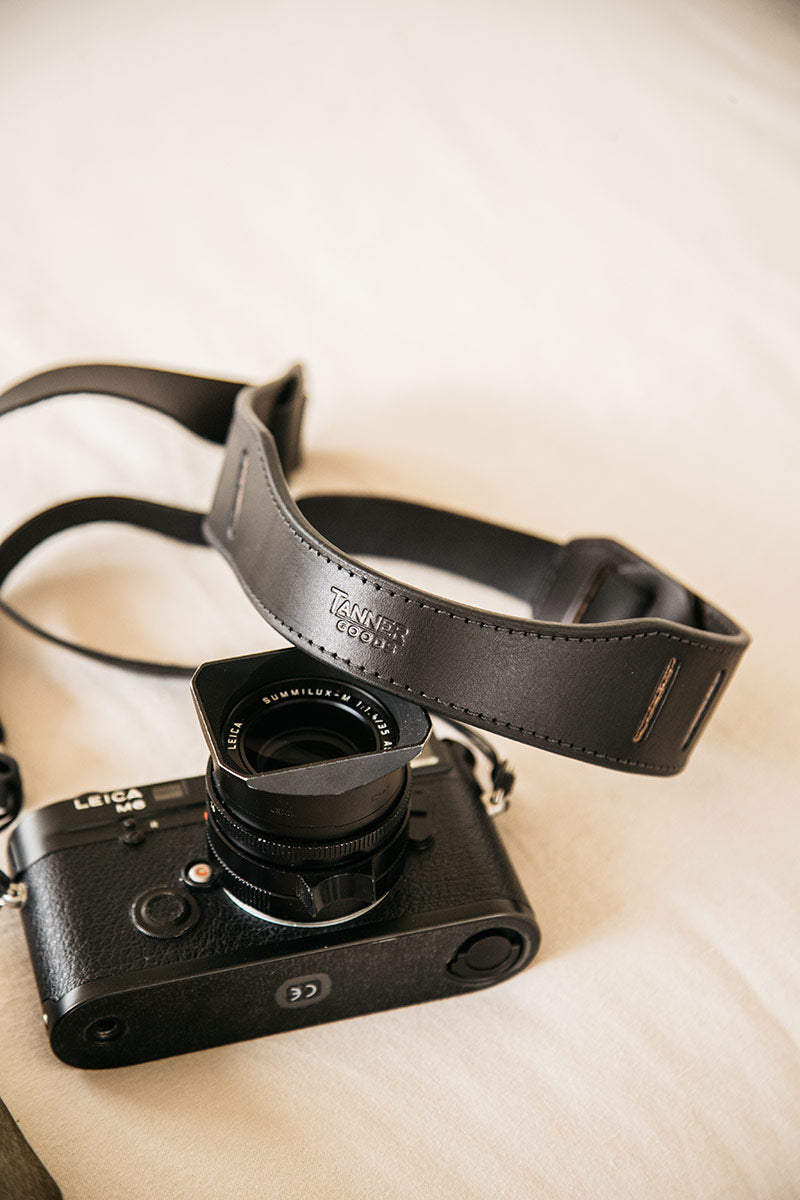 Black leather SLR camera strap from TANNER GOODS with visible stitching, on a light background next to a black camera and lens.