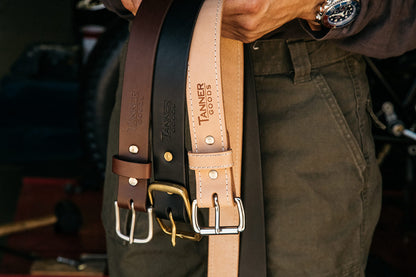 Natural leather belt displayed with brown and black leather belts, held by a person in green pants.