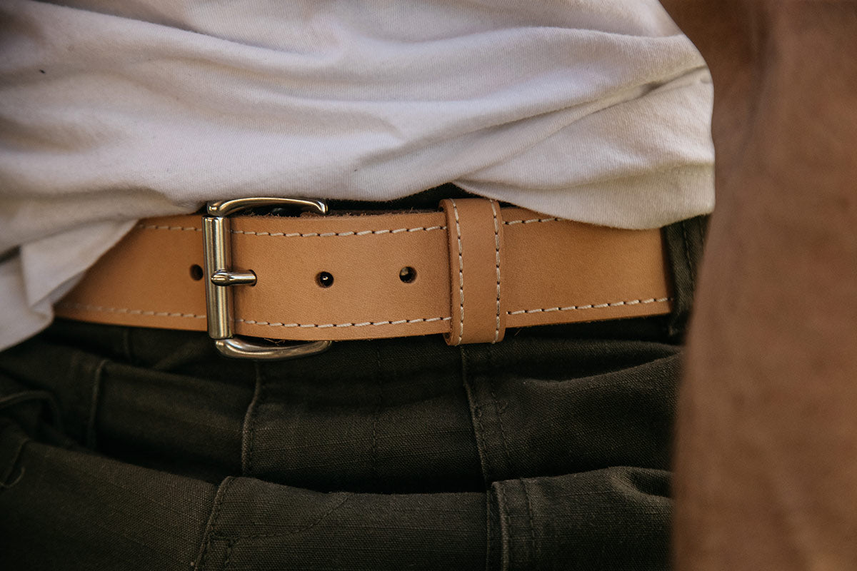 Natural leather belt with a silver buckle and white stitching, worn with a white t-shirt and dark green pants.
