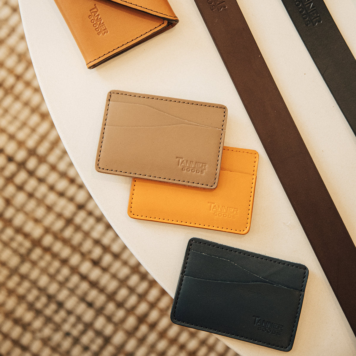 Stone grey leather wallet from Tanner Goods, displayed with orange and black cardholders and two leather belts.