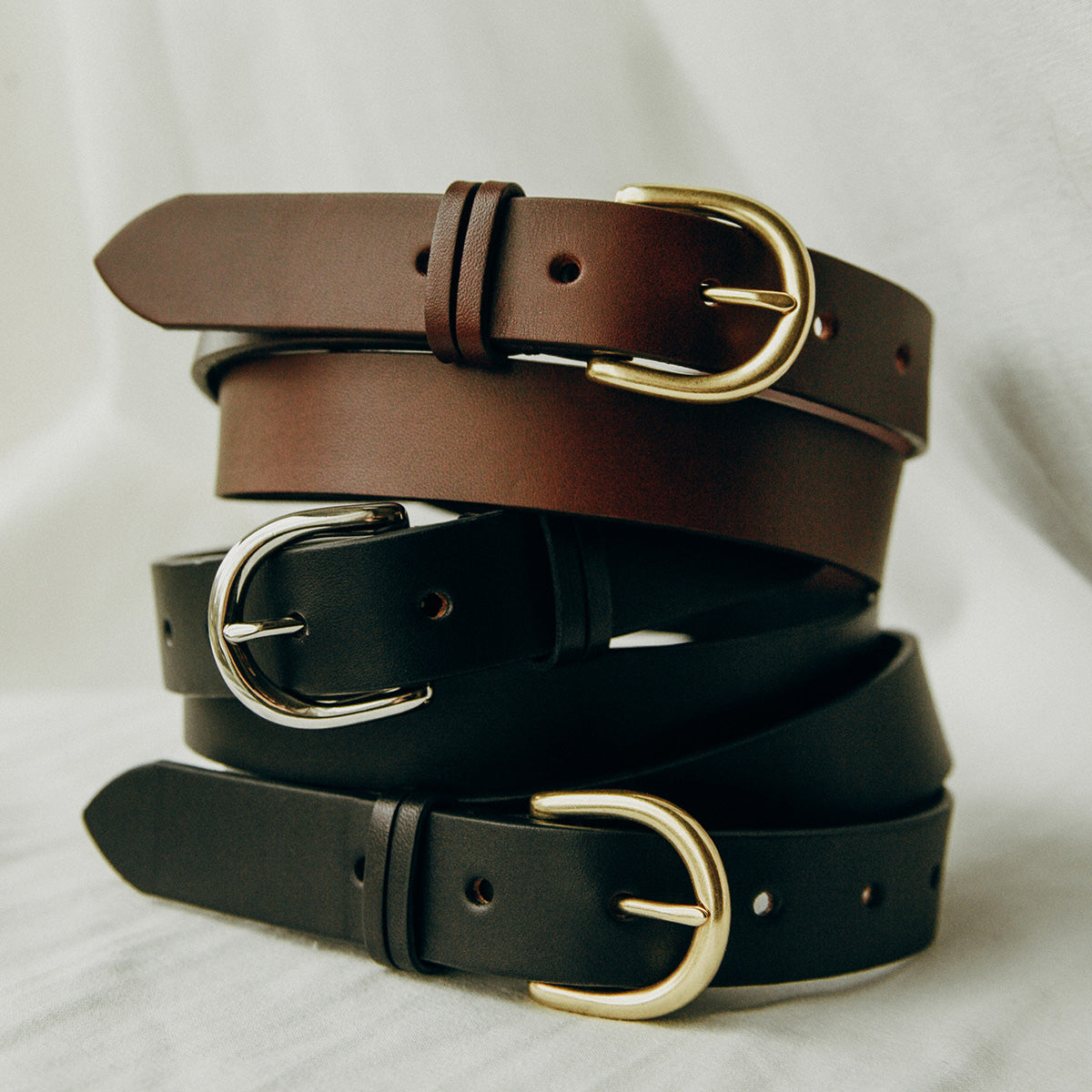 Black leather belt with a gold buckle, stacked with similar brown and black belts on a light fabric.