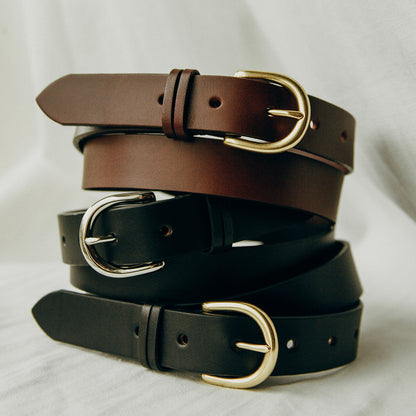 Black leather belt with a gold buckle, stacked with similar brown and black belts on a light fabric.
