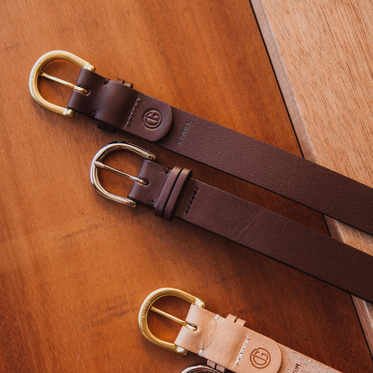 Two cognac and one natural leather Meridian Belts with brass and silver buckles, displayed on wood.