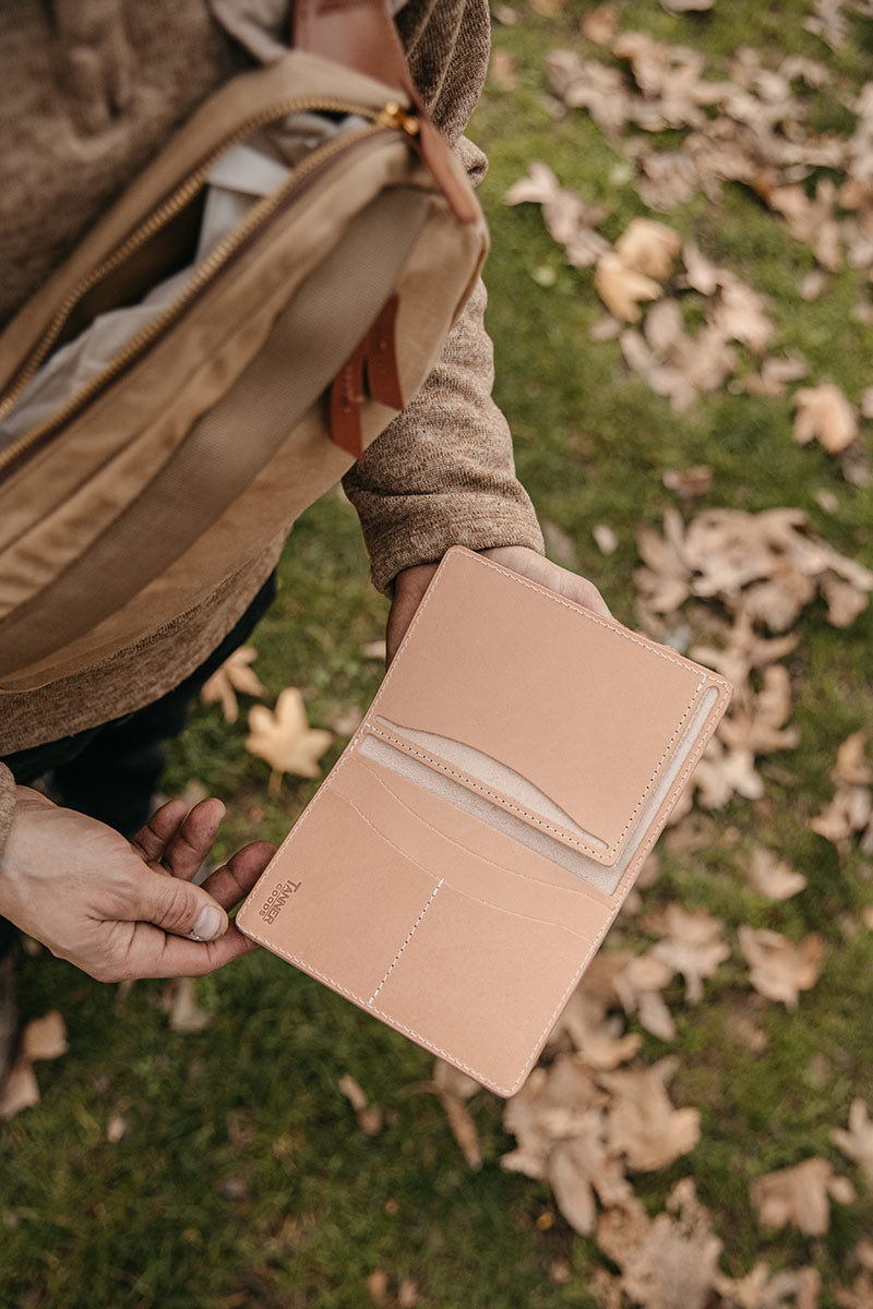 Natural leather travel wallet held open, revealing multiple card slots, by hands in a brown sweater near a tan canvas bag, over grass with autumn leaves.