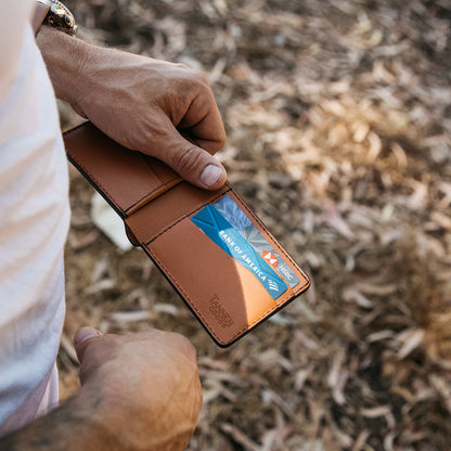 Saddle tan leather bifold wallet by TANNER GOODS, held open showing two credit cards, with a watch on the wrist.