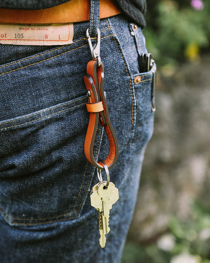 Cognac leather key lanyard hanging from a brown belt on dark blue jeans, holding two brass keys.