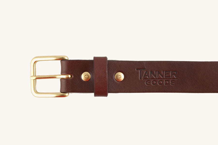 Close-up of a cognac brown leather belt by Tanner Goods featuring a gold-colored buckle, metal rivets, and stamped logo.