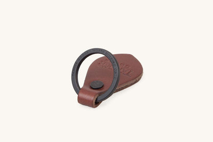 Cognac leather key fob with a black metal ring inscribed 'TANNER GOODS WORTH HOLDING ONTO', a dark rivet, and an embossed logo, angled view.