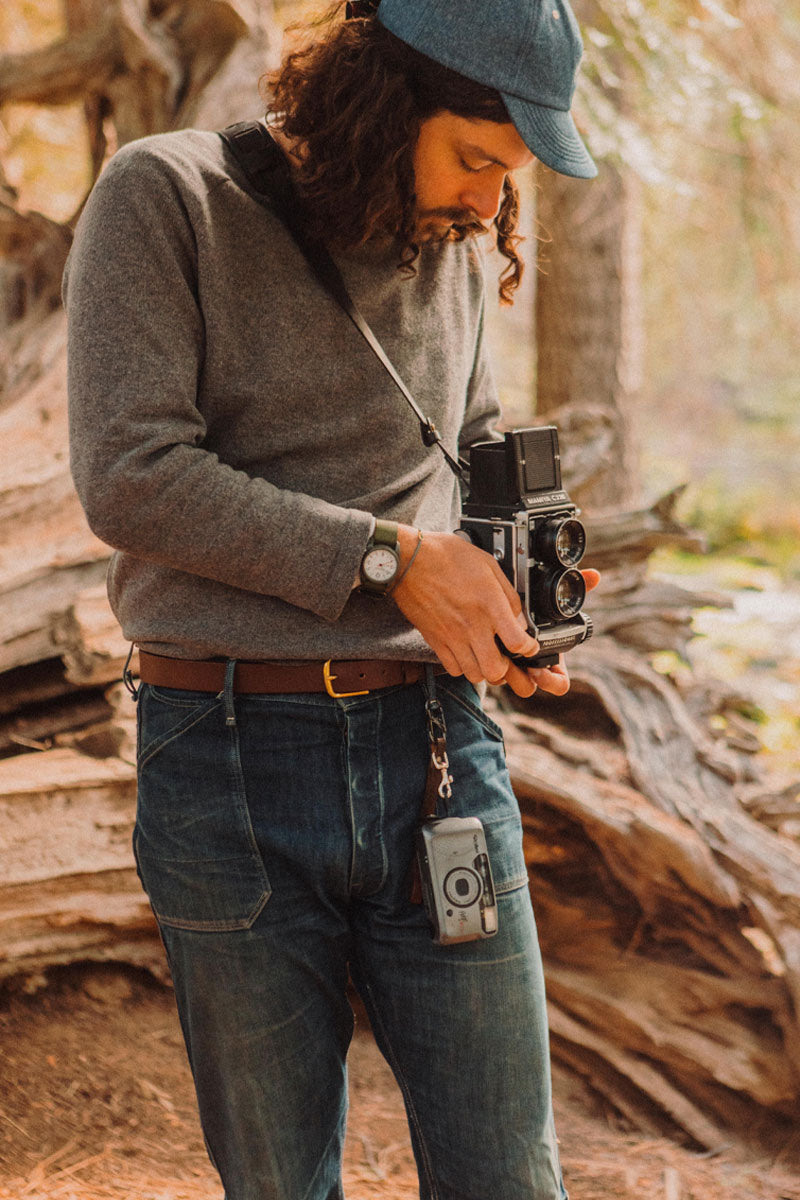Cognac leather belt with a brass buckle worn by a man in jeans and a grey sweater.