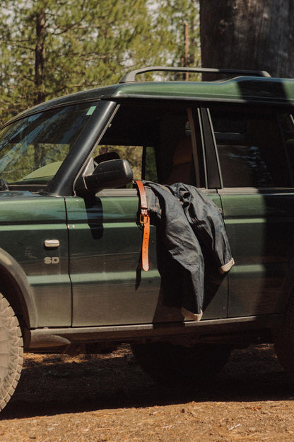 Saddle tan classic belt hanging from a dark green car door with dark blue jeans in an outdoor setting.