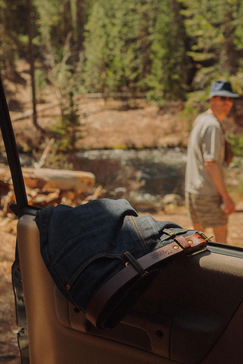Saddle tan leather belt by Tanner Goods with a brass buckle, folded over dark blue jeans in a forest setting.