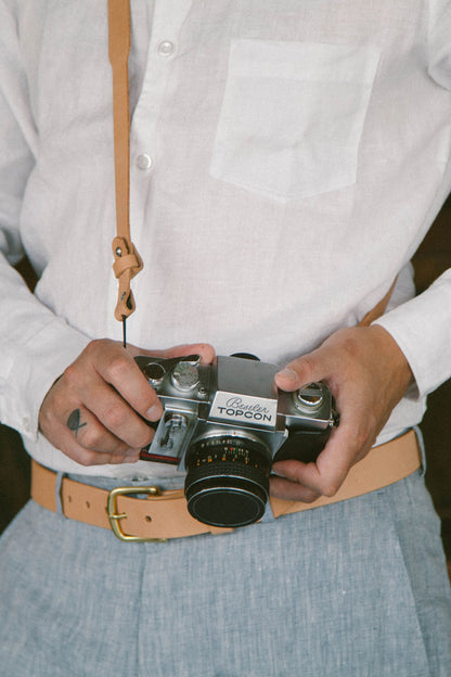 Natural leather belt with a gold-colored buckle, worn by a person in light blue trousers and a white shirt, holding a vintage camera.