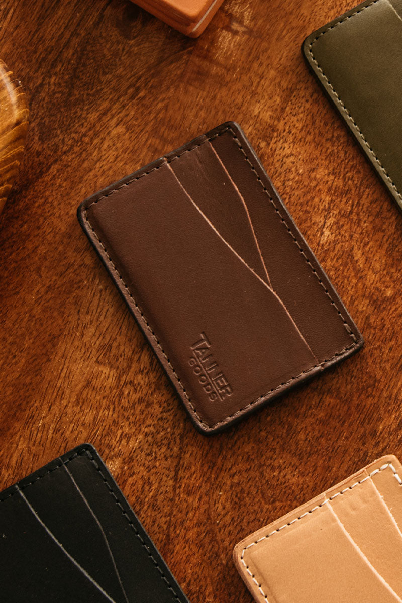Cognac brown leather wallet with 'Tanner Goods' embossed logo and stitched edges, surrounded by other wallets on wood.