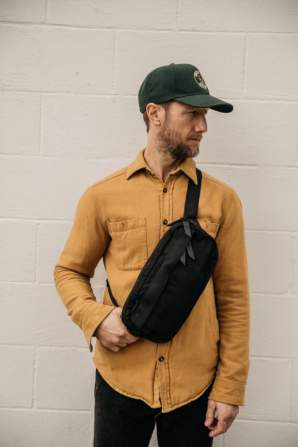 Black waxed canvas Canyon Crossbody Pack worn by a man in a yellow shirt and green cap, against a white brick wall.
