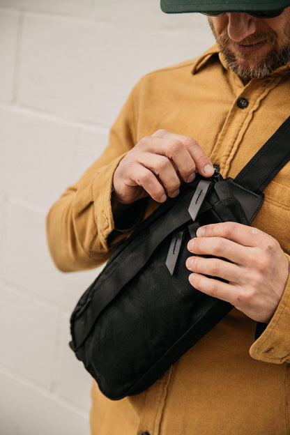 Black waxed canvas Canyon Crossbody Pack, worn by a man in a yellow shirt, being opened at the zipper.