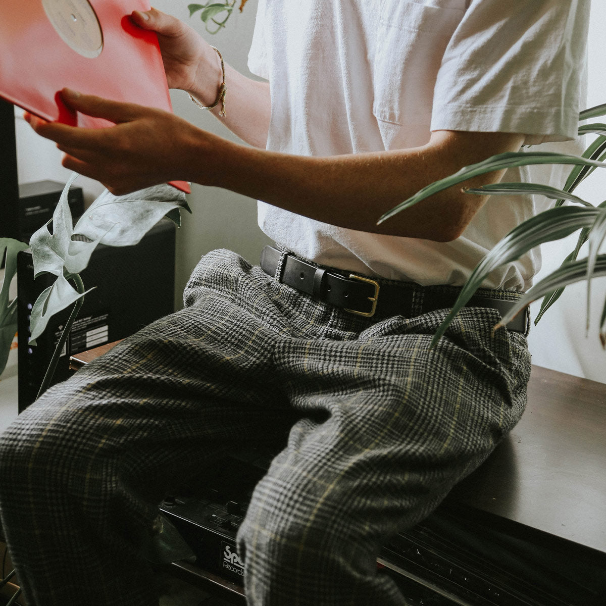 Black belt with a brass buckle, worn by a person in plaid pants holding a red vinyl record.