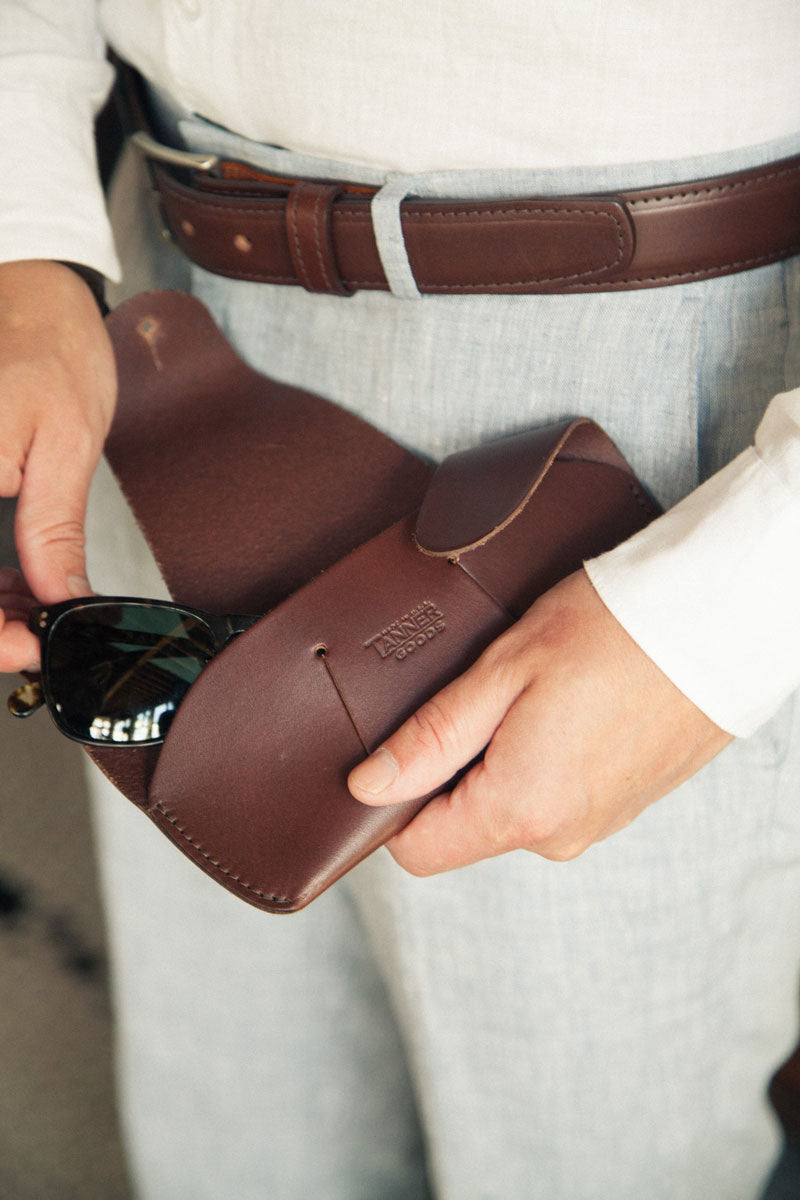 Cognac leather dress belt with a silver buckle, worn by a person in light grey pants, hands holding a brown leather sunglasses case.