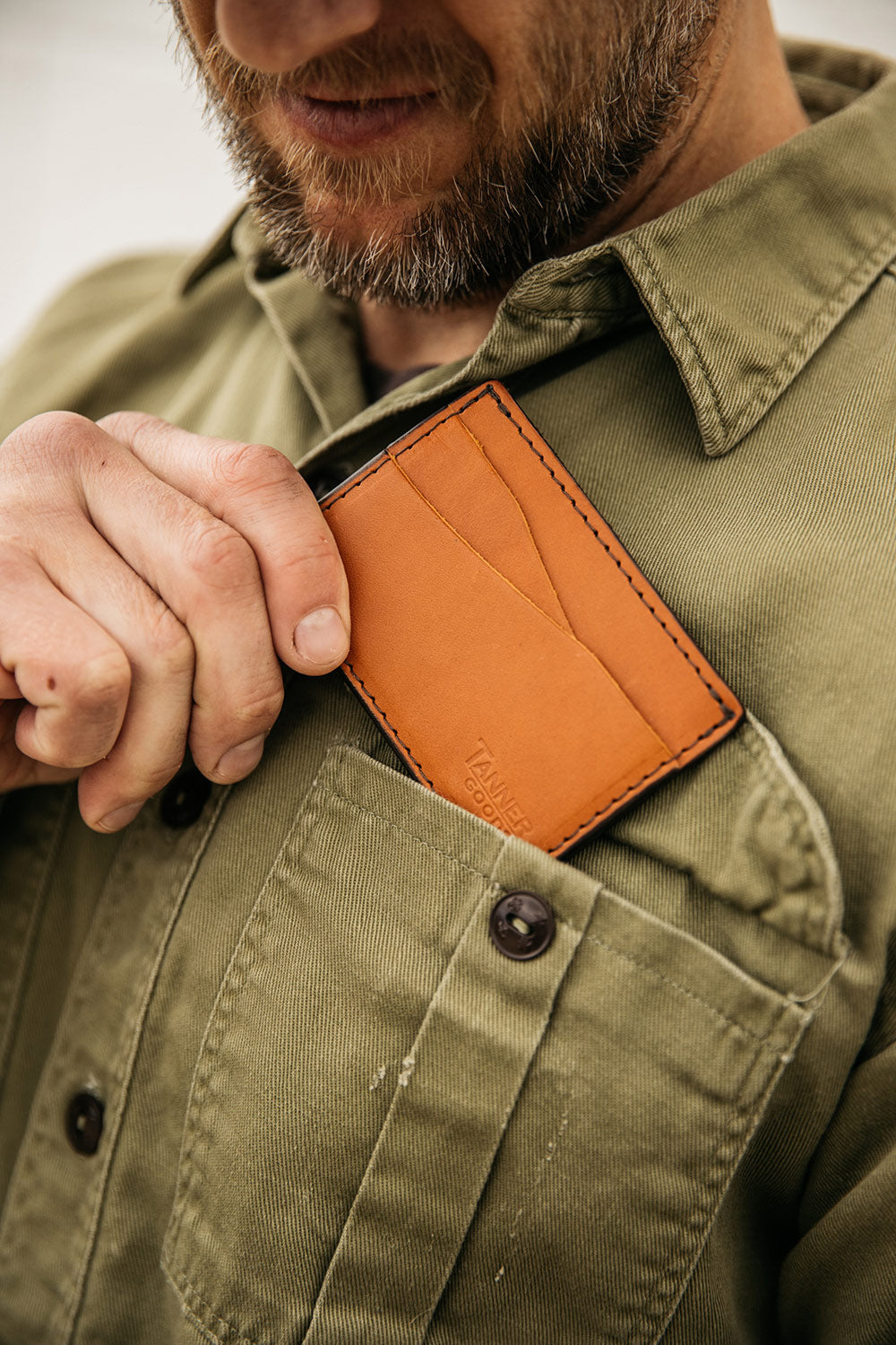 Saddle tan leather wallet by Tanner Goods being placed into a man's olive green shirt pocket.