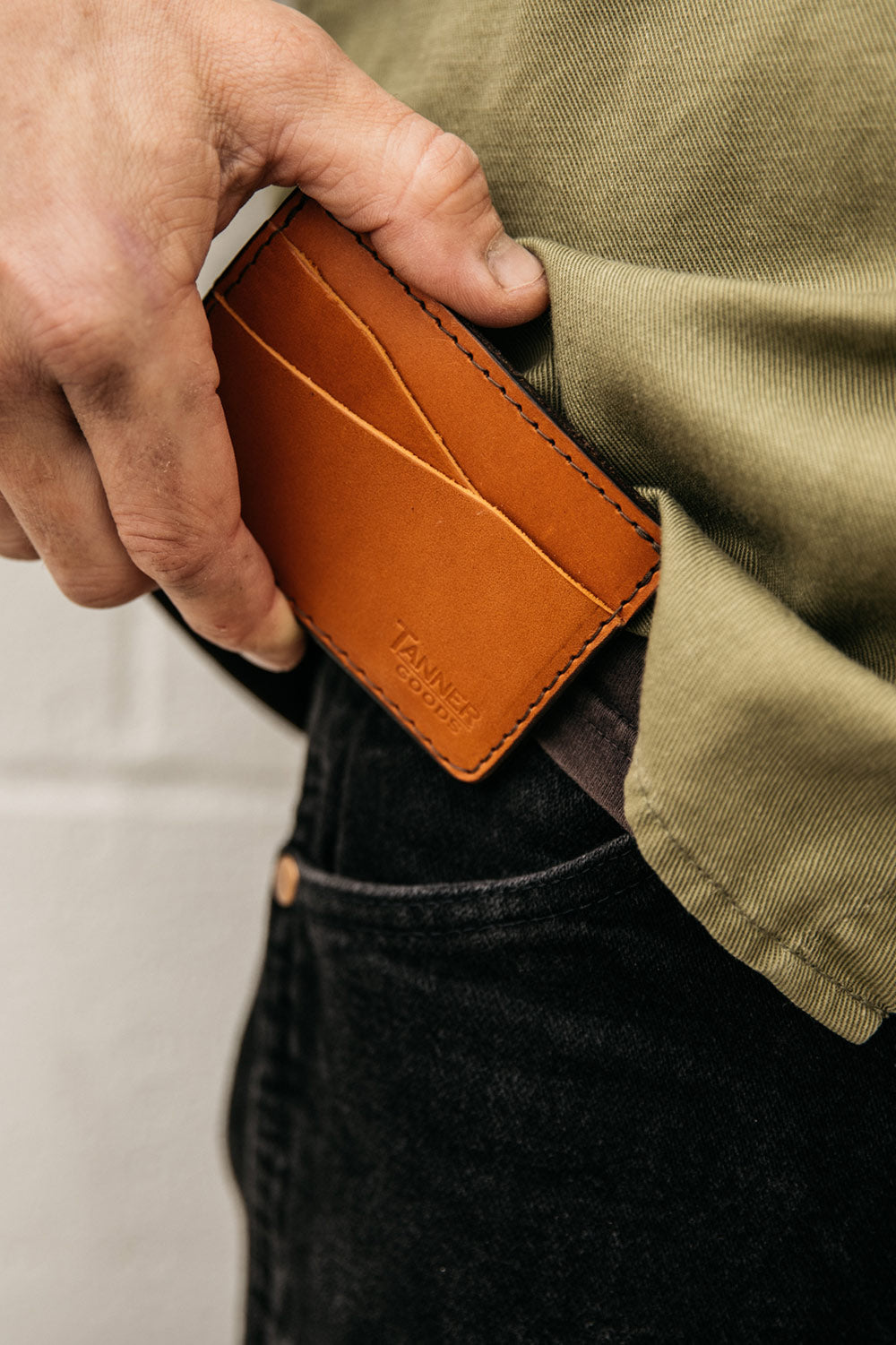 Saddle Tan leather wallet by Tanner Goods being placed into a black jeans pocket, worn with a green shirt.