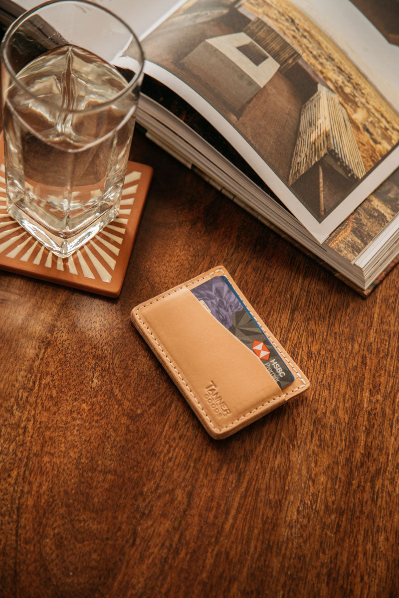 Natural leather card wallet by TANNER GOODS, with visible white stitching, holding cards on a wooden table.