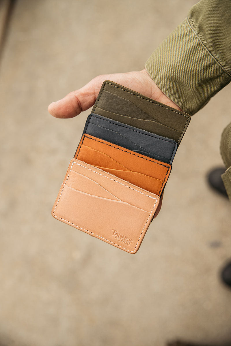 Natural leather Journeyman wallet by Tanner Goods, stacked with orange, dark gray, and olive-green leather card wallets, held in a person's hand.