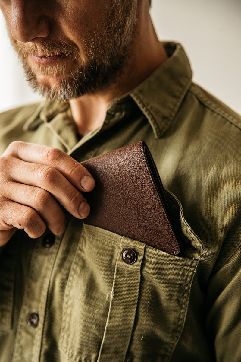 Pebbled cognac recycled leather bifold wallet being placed into the chest pocket of an olive green work shirt.