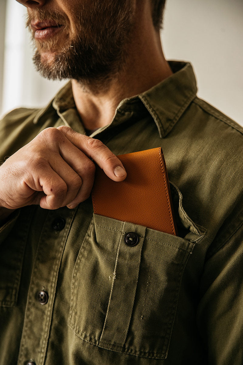 Saddle tan recycled leather bifold wallet being placed into an olive green utility shirt pocket by a man's hand.
