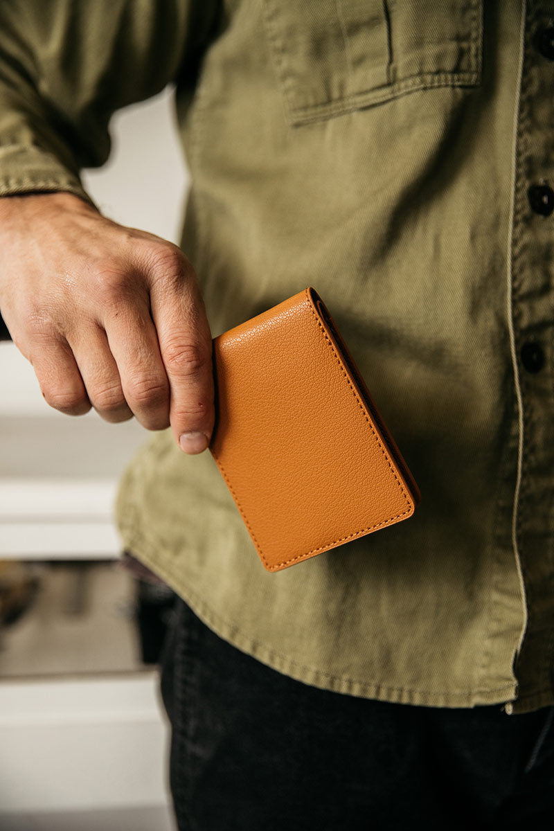 Saddle Tan recycled leather utility bifold wallet with a grained texture and visible stitching, held in a man's hand against an olive green shirt.