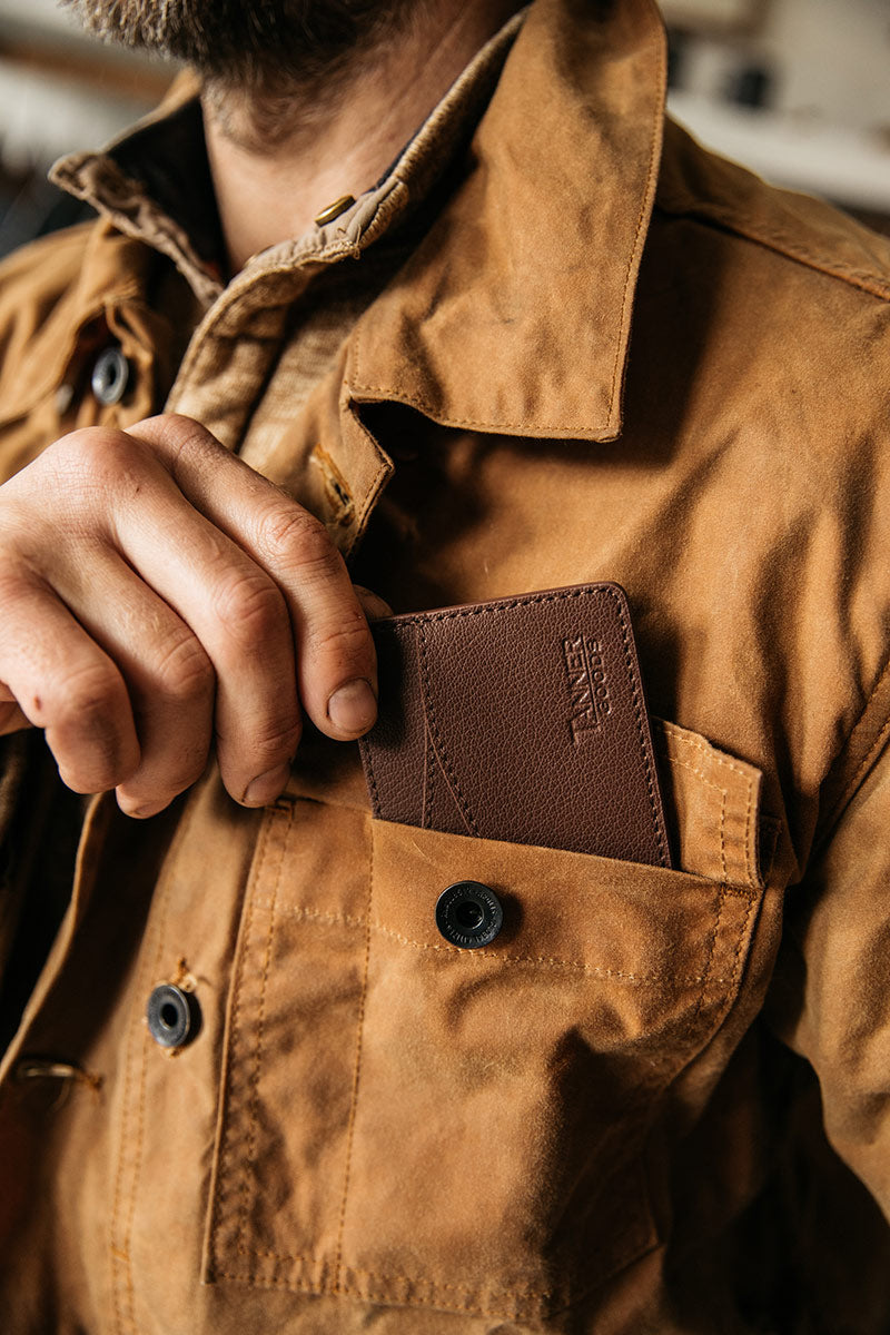 Slim cognac recycled leather wallet being placed into the chest pocket of a textured brown jacket.