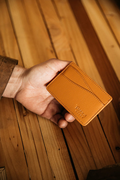 Saddle tan recycled leather wallet by TANNER GOODS, held in a hand against a rustic wooden surface, with visible card slots.