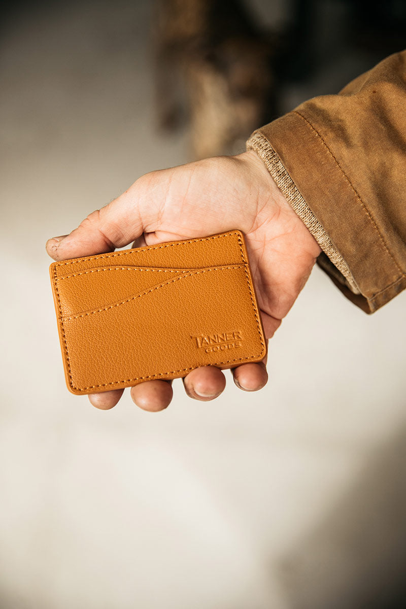 Saddle tan recycled leather wallet with curved stitch lines and embossed 'TANNER GOODS' logo, held in a hand wearing a brown jacket.