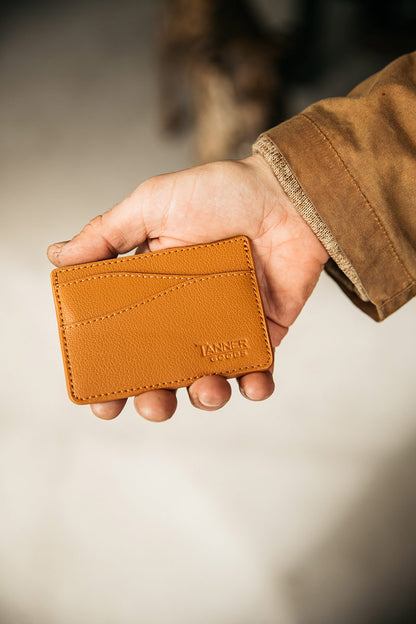 Saddle tan recycled leather wallet with curved stitch lines and embossed 'TANNER GOODS' logo, held in a hand wearing a brown jacket.