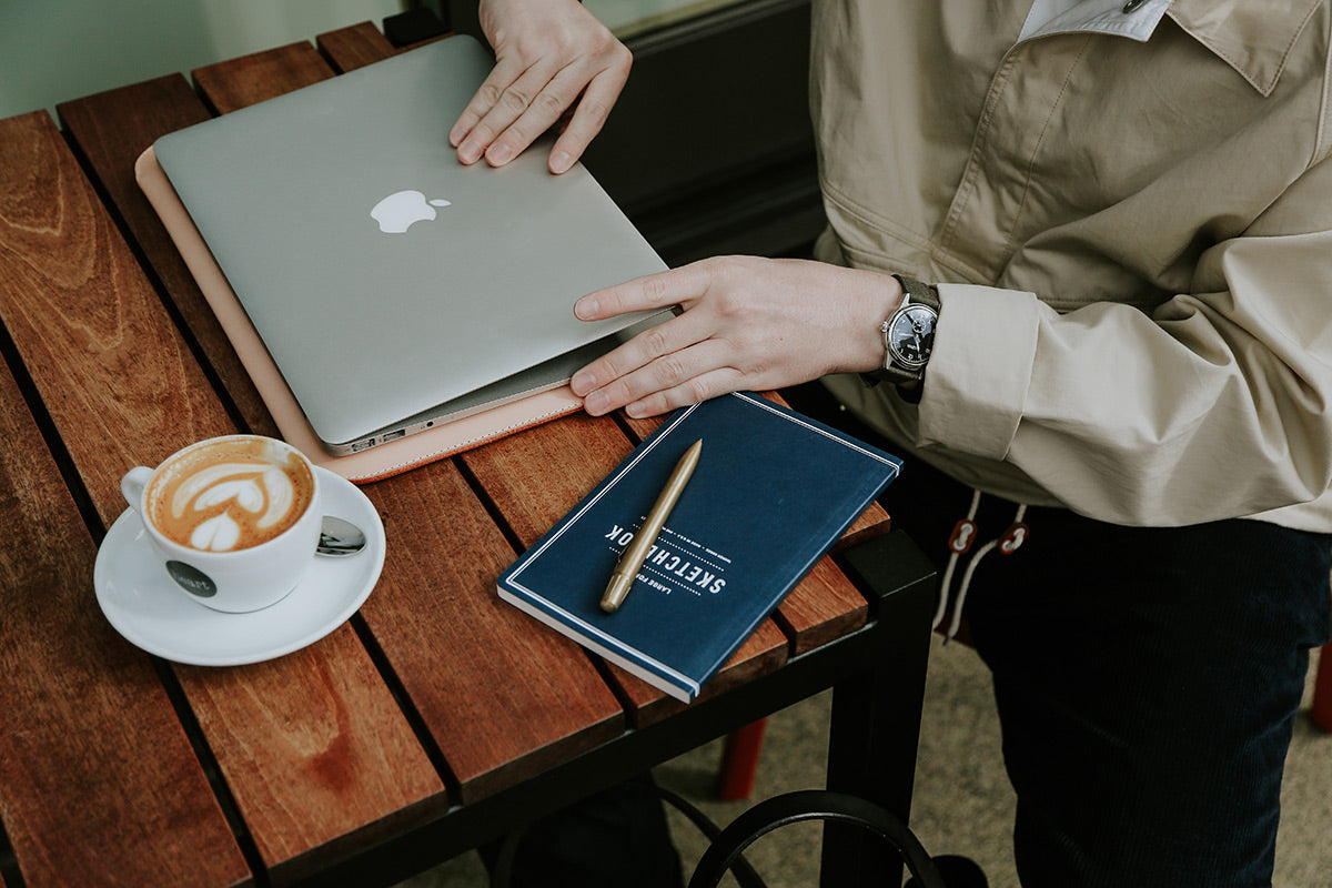 Brass pen by Memori Mechanico, lying on a blue notebook next to a silver laptop and coffee on a wooden table.