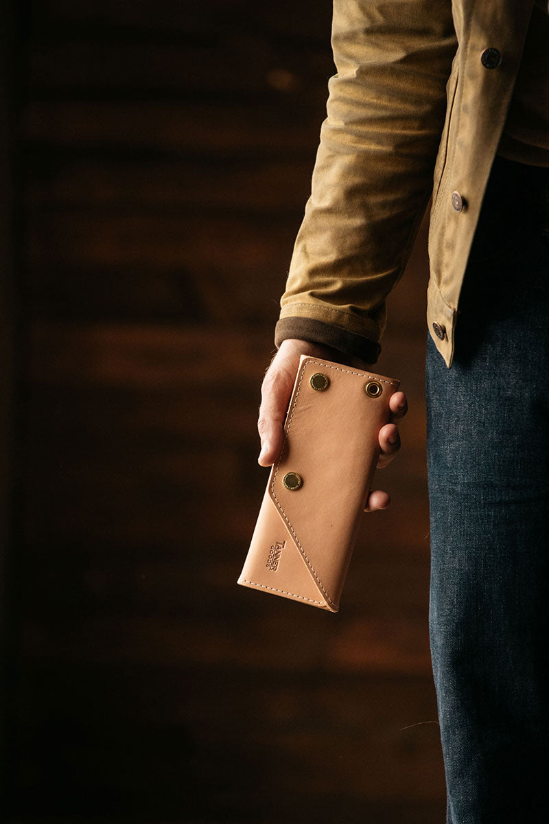 Natural leather Workman Wallet with brass snaps, held by a hand, worn with a brown jacket and blue jeans.