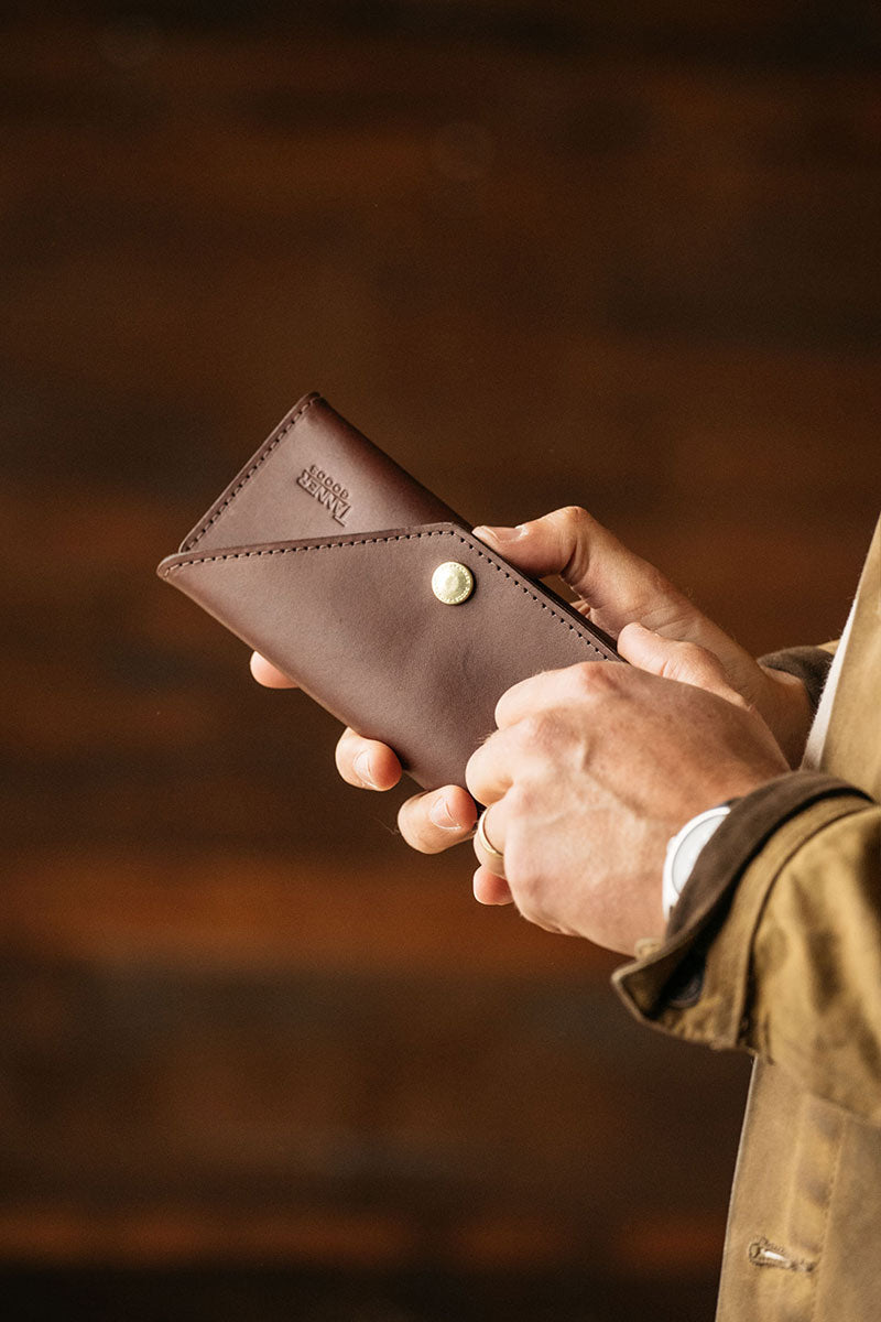 Cognac leather Workman Wallet by Tanner Goods with an envelope-style flap and brass snap closure, held open by hands.