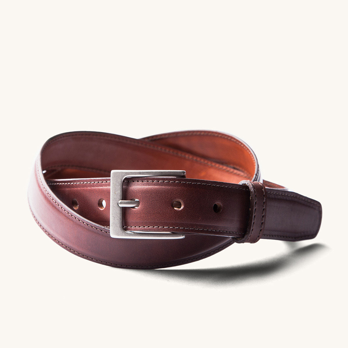 Cognac leather dress belt coiled with a silver-toned buckle and white stitching, on a white background.