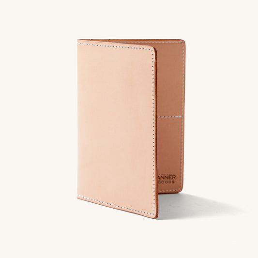 Natural leather travel wallet, standing partially open, showing neat white stitching and interior pocket with 'ANNER GOODS' text.